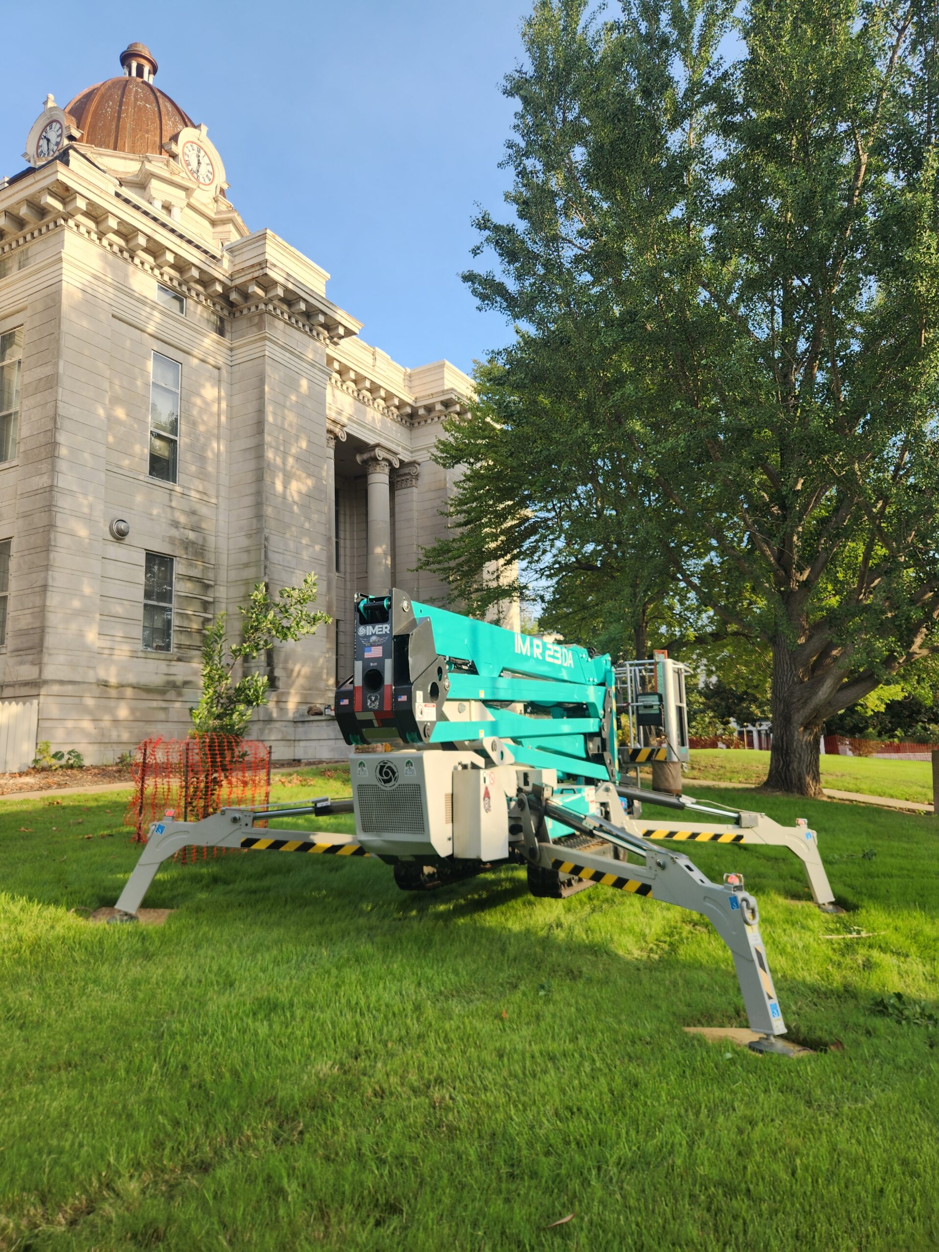 A teal IMER tracked aerial lift with extended outriggers parked on the lawn of a large stone courthouse.